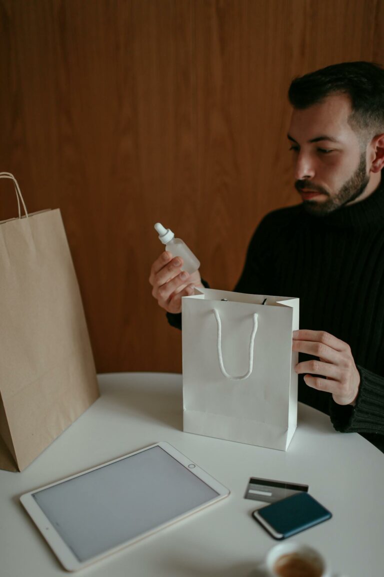 Crop self assured young bearded male buyer in casual clothes sitting at table and checking delivered purchases made via tablet with credit card at home